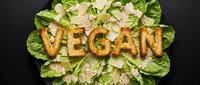Fresh vegan Caesar salad with crispy crouton letters spelling VEGAN, topped with shaved parmesan and romaine lettuce on dark background.