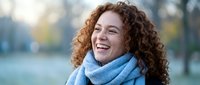 Happy young woman with curly red hair laughing outdoors in blue scarf during autumn day, expressing joy and positive emotions in natural park setting.