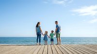 Young family with two children standing on wooden pier overlooking calm ocean waters on sunny day, ideal for travel, vacation, and lifestyle marketing materials.