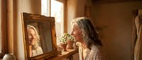 Mature woman with gray hair looking at herself in vintage mirror during morning routine in warm sunlit bedroom with natural lighting and cozy atmosphere.