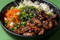 Asian beef bowl with glazed teriyaki meat, shredded carrots, bean sprouts, green onions and sesame seeds served in traditional black stone bowl on green background.