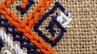 Colorful embroidered textile with orange and blue geometric patterns on natural burlap fabric background for traditional craft and needlework projects.