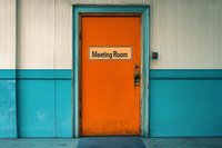 Orange meeting room door with retro sign against turquoise walls creates striking color contrast in minimalist office hallway setting, highlighting corporate workspace design.