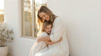 Young Caucasian mother and daughter embracing tenderly in white dresses on sunny porch, expressing love and family bonding in warm natural light for lifestyle content.