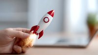 Hand holding small toy rocket with flame effect against blurred office background, symbolizing business startup launch and innovation concept for entrepreneurship growth.