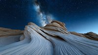 Dramatic sandstone rock formations under starry night sky with Milky Way galaxy. Natural wavy patterns in stone create surreal desert landscape at twilight.