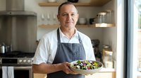 Middle-aged Caucasian male chef wearing white shirt and gray apron presenting fresh salad in modern kitchen with natural light and contemporary design.