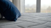 Close-up of blue pillow on gray quilted mattress near window with blurred cityscape view, suitable for bedding, comfort, and home interior design projects.