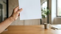 Male hand holding blank white paper sheet at wooden desk with office supplies and blurred background, suitable for mockups, presentations, and business communications.