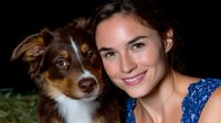 Young Caucasian woman with brown hair in blue dress smiling while posing with Australian shepherd mix dog against dark background, showing bond between pet and owner.