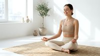 Young woman of mixed ethnicity in lotus position on beige mat in bright minimalist room with potted plant, practicing meditation and mindfulness for wellness and mental health.