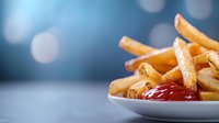 Golden crispy french fries served on white plate with red ketchup sauce, close-up view against blurred blue bokeh background. Fast food restaurant menu.