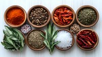 Aromatic herbs, spices and seasonings in clay bowls on white wooden background, including paprika, black pepper, dried chili peppers, fresh sage leaves and sea salt.