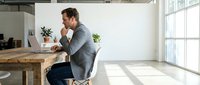 Young professional man working on laptop at wooden desk in bright modern office space with natural light streaming through large windows.