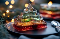 Glass Christmas tree shaped dessert with layered colorful jelly on dark plate against blurred holiday lights background, festive winter table setting.