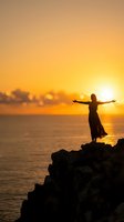 Woman silhouette with open arms standing on rocky cliff overlooking ocean at golden sunset for freedom and wellness concepts.