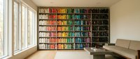 Modern home library with color-coordinated bookshelf displaying rainbow arrangement of books in contemporary living room with natural light and seating area.