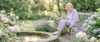 Senior Caucasian woman in lavender outfit relaxing on wooden garden bench surrounded by blooming flowers and lush greenery in peaceful park setting.