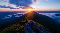 Dramatic mountain peak at sunrise with sun rays breaking through clouds, aerial view of misty mountain ridge surrounded by low-lying fog in morning light.