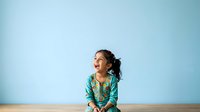 Young girl with dark hair and medium skin tone wearing a teal patterned shirt, sitting against a blue wall background, looking upward with a thoughtful expression, suitable for educational, lifestyle, and children's content.