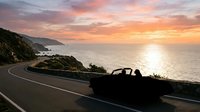 Silhouette of convertible car driving on coastal road at sunset with ocean view and dramatic sky, suitable for travel and automotive marketing.