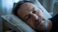 Middle-aged man with dark hair and facial stubble sleeping peacefully on a gray linen pillow, eyes closed, indoors during daytime with soft natural lighting.