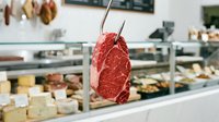 Raw red meat steak held on fork in butcher shop with display case of fresh cuts and products visible in background.