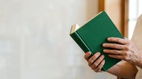 Person holding open green hardcover book with gold-trimmed pages near window with natural light, suitable for education, literature, reading, and knowledge-related projects.