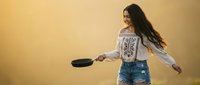 Young Hispanic woman in embroidered white blouse and denim shorts dancing with cast iron skillet in kitchen, celebrating cooking and culinary joy.