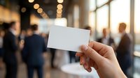 Hand holding blank white business card in modern office environment with blurred professional people in background for networking and corporate branding.