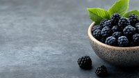 Fresh ripe blackberries in ceramic bowl with green leaves on dark stone background, copy space for text. Food photography styling.