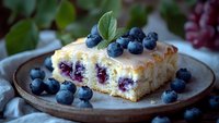 Fresh blueberry cake slice with vanilla glaze and fresh berries on rustic ceramic plate, soft natural lighting creates appetizing dessert atmosphere.