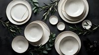 Overhead view of white ceramic dinnerware set with stacked plates and bowls arranged on dark surface with fresh green foliage and small bowls for table setting and dining design.