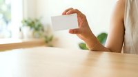 Man with medium skin tone holding blank white business card at wooden table indoors, suitable for corporate branding, marketing materials, and professional networking concepts.