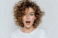 Young Caucasian woman with curly brown hair showing surprised expression with open mouth wearing white shirt on gray background for lifestyle content.
