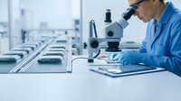 Laboratory technician in blue coat and protective gloves examining samples under microscope in modern research facility with equipment rows in background.