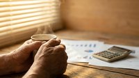 Senior adult hands holding eyeglasses over financial documents and calculator on wooden desk with warm sunlight streaming through blinds for retirement planning and vision care concepts.