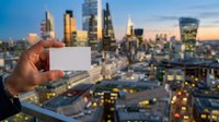 Caucasian male hand holding blank white business card against illuminated urban skyline at dusk, suitable for corporate branding, networking concepts, and business marketing materials.