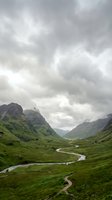 Scenic mountain valley with winding river through lush green meadows, dramatic cloudy sky, and rugged peaks, suitable for travel and nature backgrounds.