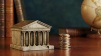 Miniature classical building model with columns next to stacked coins on wooden desk with books and globe, representing finance, banking, investment concepts.