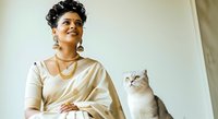 Young woman with dark curly hair and South Asian ethnicity wearing beige linen shirt holding gray and white cat indoors. Pet owner portrait.