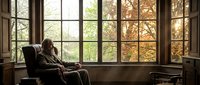 Elderly man relaxing in leather armchair by large windows overlooking autumn forest. Peaceful retirement lifestyle in cozy home interior with warm natural lighting.