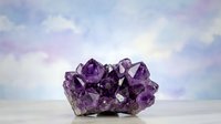 Purple amethyst crystal cluster on white surface against soft pastel sky background for healing meditation and spiritual wellness concepts.