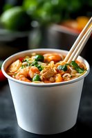 Spicy Asian noodle soup with shrimp, chicken, green onions and peas in white takeout container with wooden chopsticks for food delivery service.
