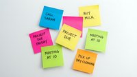 Colorful sticky notes with handwritten reminders and tasks scattered on white surface, including calls, shopping, meetings, and project deadlines for organization and planning.