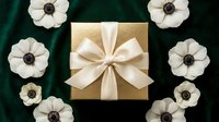Elegant gold gift box with cream ribbon surrounded by white anemone flowers on emerald green velvet background for luxury holiday celebrations.