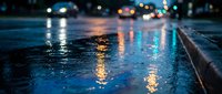 Wet asphalt street reflecting colorful city lights and car headlights at night creating atmospheric urban scene with bokeh effect and water puddles on road surface.