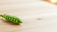 Fresh green pea pod with visible peas inside on wooden cutting board surface for healthy cooking and organic vegetable preparation concepts.