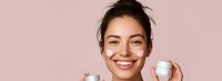 Young woman with radiant smile applying facial cream on cheeks, holding skincare product jar against soft pink background for beauty and cosmetic advertising.
