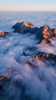 Dramatic mountain peaks emerging through sea of clouds at sunrise with golden light illuminating rocky summits above misty valley fog creating ethereal landscape.